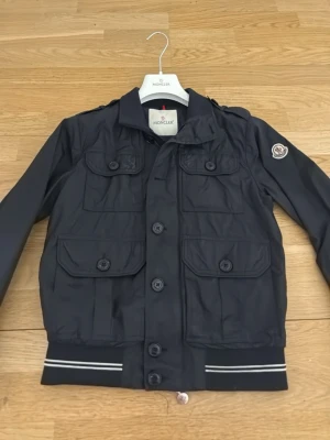 Svart Moncler jacka med fickor - Snygg mörkblå jacka från Moncler med fyra stora fickor framtill och ribbade muddar med vita ränder. Jackan har knappstängning och en klassisk Moncler-logga på ärmen. Perfekt till våren och sommaren storlek 0 pris kan diskuteras👍