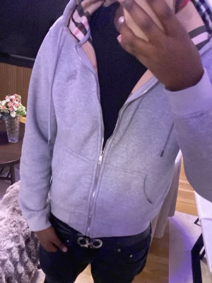 Grå zip hoodie med Burberry-mönster - Snygg grå hoodie med dragkedja och huva från Burberry. Utsidan är enkel och stilren medan insidan har det klassiska Burberry-rutiga mönstret i beige, svart och rött. Hoodien har fickor framtill och ribbade muddar. Perfekt för dig som vill ha en exklusiv streetwear-look.