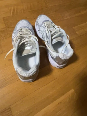 Vita och silver Asics sneakers - Säljer ett par fräscha Asics sneakers i vitt och silver. Skorna har mesh- och syntetmaterial för bra andningsförmåga och komfort. Klassisk låg modell med snörning och tydliga Asics-loggor på sidorna. Perfekta för dig som gillar sportig och clean stil.