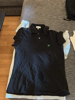 Svart pikétröja från Lyle & Scott - Klassisk svart pikétröja från Lyle & Scott med krage, knappar framtill och den ikoniska gröna loggan broderad på bröstet. Tröjan har korta ärmar och är tillverkad i mjuk bomull som känns skön mot huden.