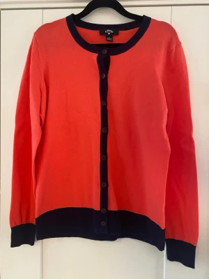 Röd kofta med svarta detaljer från Callaway strl M - Snygg röd kofta från Calloway med marinblå kontrastdetaljer vid krage, knappslå, ärmslut och nederkant. Rund halsringning och knäppning framtill med svarta knappar. Perfekt för lager på lager och enkel att styla till olika outfits.  Nya, aldrig använd   Mått Över bröstet: ca 52 cm Längd från axeln och ner: ca 64 cm  70% bomull och 30% nylon  Kommer från ett djur och rökfritt hem