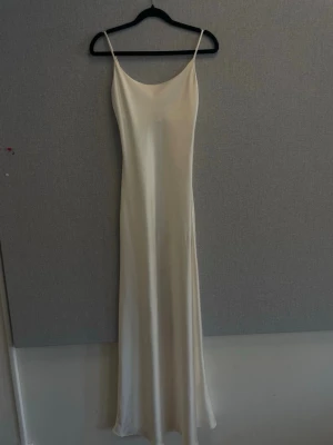 Lång vit satinklänning med tunna band - Elegant långklänning i vit satin med smala axelband och enkel, stilren siluett. Klänningen har en mjukt rundad urringning och faller snyggt längs kroppen. Perfekt för dig som gillar minimalistisk och tidlös stil.