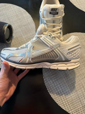 Nike Vomero 5 vita och silver sneakers - Nike Vomero 5 sneakers i vitt och silver med mesh- och syntetpaneler. Skorna har en chunky sula, reflekterande detaljer och klassisk swoosh på sidan. Perfekta för dig som gillar sportig och trendig stil. Skön passform och snygg retrovibe.