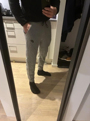 Gråa mjukisbyxor från Nike - Säljer ett par gråa mjukisbyxor från Nike med svart logga på vänster lår. Byxorna har dragsko i midjan, muddar vid bensluten och två sidofickor. Tillverkade i mjukt och bekvämt material, perfekta för chill dagar eller träning.