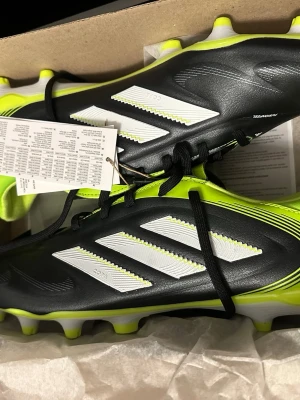 Svarta och neongula Adidas fotbollsskor - Säljer ett par snygga fotbollsskor från Adidas i svart och neongul med klassiska vita ränder. Skorna har snörning och är tillverkade i syntetmaterial, perfekta för spel på gräsplan. Modern design som sticker ut på planen. Helt ny inte använd köpte den men de var inte rätt storlek.