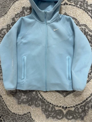 Ljusblå Nike NOCTA hoodie - Snygg ljusblå hoodie från Nike NOCTA med dragkedja framtill och två fickor med dragkedja. Hoodien har en klassisk huva och diskret Nike-logga på bröstet samt NOCTA-tryck på ärmen. Tillverkad i mjukt material som känns skönt mot huden.