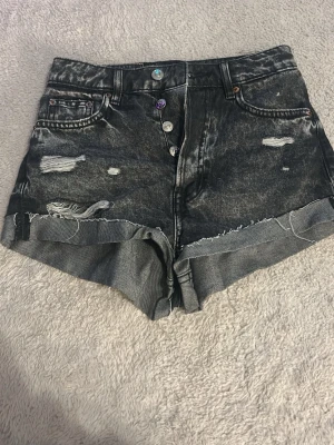 Svarta denimshorts med slitningar - Säljer ett par svarta denimshorts med slitna detaljer och råa kanter. Shortsen har hög midja, knappar framtill och klassiska fem fickor. Perfekta för en avslappnad och trendig look.
