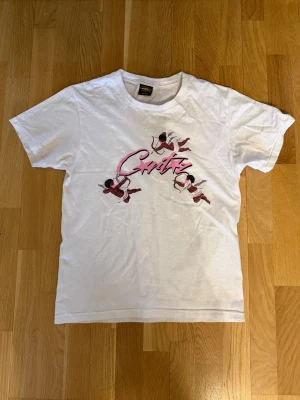 Vit Critiz t-shirt med tryck - Vit t-shirt från Critiz med korta ärmar och ett färgglatt tryck framtill som visar tre tecknade änglar och texten 'Corteiz' i rosa. Klassisk passform och rund halsringning. Perfekt för dig som gillar streetwear och vill sticka ut lite extra.