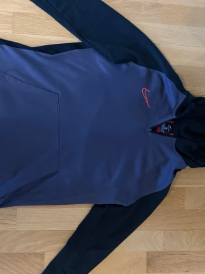 Nike hoodie i svart och mörkgrå - Nike hoodie i svart och mörkgrå med röd logga på bröstet. Klassisk huva och stor magficka framtill. Tillverkad i mjukt och bekvämt material, perfekt för chill dagar. Snygg kontrast mellan ärmar och kropp. Halvlång dragkedja vid halsen.