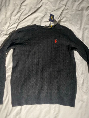 Svart kabelstickad tröja Polo Ralph Lauren - Kabelstickad svart tröja från Polo Ralph Lauren med rund halsringning och den klassiska röda logotypen broderad på bröstet. Tröjan har långa ärmar och ribbade muddar vid ärmslut och nederkant. Perfekt för en stilren och tidlös look.
