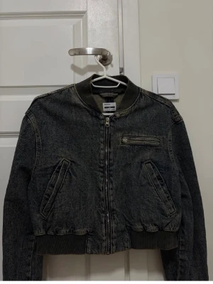 Vintage jacka  - Snygg mörkgrå jeansjacka i bombermodell. Jackan har dragkedja framtill, ribbade muddar vid ärmslut och midja samt tre fickor, varav en med dragkedja på bröstet. Perfekt för en cool och avslappnad stil. Den har en gammaldags stil och är typ vintage 