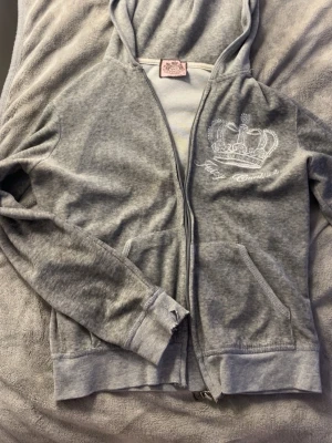 Grå hoodie från Juicy Couture - Grå hoodie från Juicy Couture med dragkedja, huva och fickor framtill. På bröstet finns ett broderat kronmotiv med strassdetaljer och Juicy Couture-loggan. Mjuk och bekväm, perfekt för en avslappnad look. Men har ribbade muddar och tappat några strass stenar 