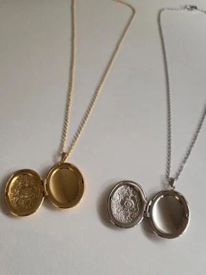 Ovala medaljonghalsband i guld och silver - Två ovala medaljonghalsband, ett i guldfärg och ett i silverfärg. Båda har ett vackert blommönster på framsidan och kan öppnas för att lägga in små bilder eller minnen. Kedjorna är tunna och eleganta i rostfritt stål, perfekta för att ge en personlig touch till din stil.