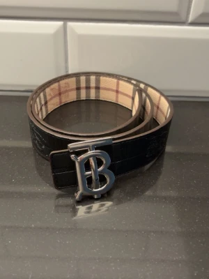 Burberry bälte med TB-spänne - Snyggt Burberry bälte med klassiskt rutigt mönster på insidan och ett stort TB-spänne i silver. Utsidan är svart och bältet är gjort i skinn. Perfekt accessoar för att lyfta din outfit.