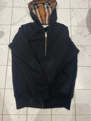 Svart hoodie från Burberry med dragkedja - Säljer en svart hoodie från Burberry med dragkedja framtill och klassiskt rutigt foder i huvan. Hoodien har fickor på sidorna, ribbade muddar och snören i huvan. Perfekt för dig som gillar stilrena plagg med en twist från ett ikoniskt märke.