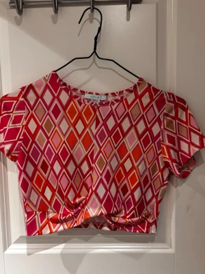 Färgglad croppad topp från Amisu - Säljer en croppad topp från Amisu med korta ärmar och ett färgstarkt rutmönster i rosa, rött, orange och beige. Toppen har twistad detalj framtill och är tillverkad i ett mjukt, glansigt material som känns skönt mot huden. Perfekt för dig som gillar statement-plagg!