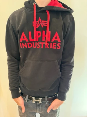 Röd Svart Alpha Industries Hoodie - Säljer en för fet röd svart Alpha Industries hoodie i storlek S. Modellen på bilden är 180 cm lång och väger 65 kg.
