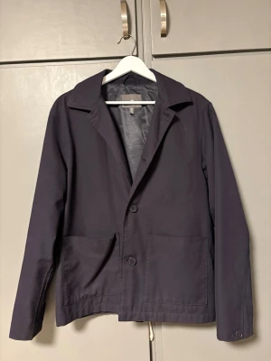 Mörkblå overshirt från H&M - Stilren mörkblå overshirt från H&M med två stora framfickor och klassisk krage. Jackan har knäppning med två knappar framtill och är tillverkad i ett mjukt bomullsmaterial. Perfekt för dig som gillar en clean och enkel look.