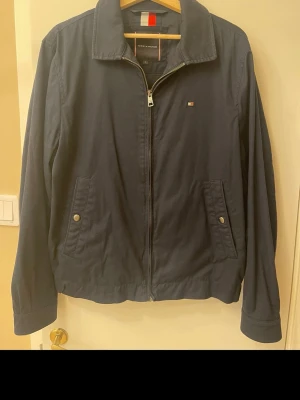 Marinblå vindjacka Tommy Hilfiger - Snygg marinblå vindjacka från Tommy Hilfiger med dragkedja framtill, två fickor med tryckknappar och klassisk krage. Liten logga på bröstet och diskreta detaljer. Perfekt för dig som vill ha en stilren och enkel jacka till vardags.