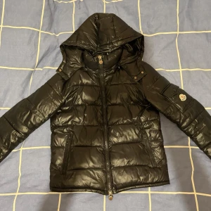 Svart dunjacka från Moncler med huva - Snygg svart dunjacka från Moncler med avtagbar huva och dragkedja framtill. Jackan har två stora fickor med dragkedja och Moncler-logga på ärmen. Perfekt för kalla vinterdagar och riktigt stilren look. Priset är inte spikat.