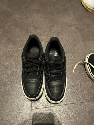 Svarta Nike Air Force 1 sneakers - Klassiska svarta Nike Air Force 1 sneakers i lågt utförande med svart läder, svarta skosnören och vit sula. Swoosh-logga på sidan och AIR-detalj på sulan. Perfekta för dig som gillar en clean och tidlös stil.