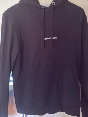 Svart hoodie Jack n Jones - Svart hoodie från Jack n Jones med broderad vit text på bröstet. Klassisk modell med huva, dragsnören och stor magficka framtill. Tillverkad i mjukt bomullsmaterial och har ribbade muddar vid ärmslut och nederkant. Perfekt för en chill och avslappnad stil.