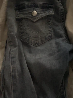 Blå jeans från True Religion - Säljer ett par klassiska blå jeans från True Religion med ikonisk fickdesign och silverfärgade knappar. Jeansen har låg midja, dom är flare och detaljerade sömmar på bakfickan. Perfekta för en avslappnad och trendig look.