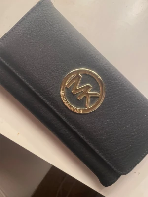 Michael Kors plånbok - Stilren svart plånbok från Michael Kors i skinn med guldfärgad MK-logga på framsidan. Klassisk rektangulär form och enkel design som ger en lyxig känsla. Perfekt för att hålla ordning på kort och kontanter.