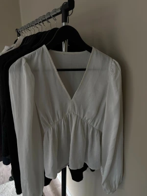 Vit blus med v-ringning och puffärm - Säljer en vit blus med djup v-ringning och långa puffärmar. Blusen har en lös, flowy passform med rynk under bysten för en feminin siluett. Tillverkad i ett tunt, lätt tyg som känns luftigt och skönt. Perfekt att styla med jeans eller kjol.