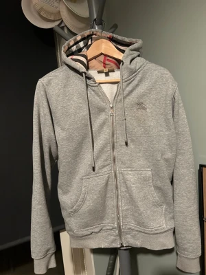 Grå zip hoodie från Burberry - Säljer en grå hoodie från Burberry med dragkedja, Den är i storlek M men passar perfekt som S dragkedjan kan vara lite krånglig ibland men det är enkelt att fixa👍
