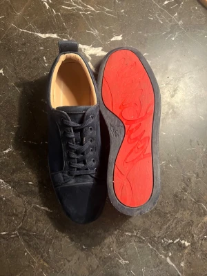 Mörkblå sneakers från Christian Louboutin - Snygga mörkblå sneakers i mocka från Christian Louboutin med klassisk röd sula och diskret logga. Skorna har rund tå, mörk blå  skosnören och låg profil. Perfekta för dig som vill ha en stilren men ändå lyxig sneaker med ikonisk detalj. 