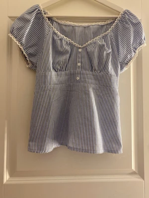 Blårandig blus med spetsdetaljer - Söt blus med blå och vita smala ränder, korta puffärmar och dekorativa spetsdetaljer längs halsringning och ärmslut. Knäppning framtill med små vita knappar och lätt rynkad midja för en feminin siluett. Perfekt till jeans eller kjol.säljer pga att den tyvärr inte kommer till användning💞