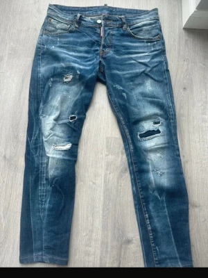 Dsqaured2 - Säljer ett par blå jeans med slitningar och hål på benen. Jeansen har fem fickor, knappgylf och en klassisk rak passform. Snyggt tvättade detaljer och en cool, avslappnad look. Perfekta för dig som gillar streetstyle och en edgy vibe.