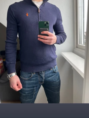 Ralph Lauren halvzip  - Snygg marinblå tröja med halv dragkedja från Polo Ralph Lauren. Klassisk orange logga på bröstet och ribbade muddar vid ärmslut och nederkant. Perfekt till jeans eller chinos för en clean och stilren look.