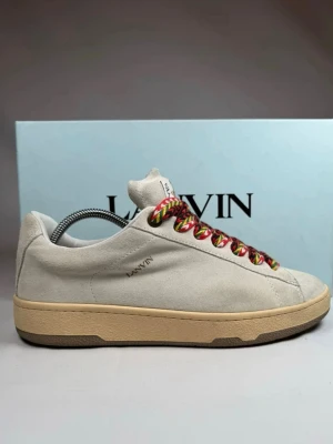 Lanvin crub lite - Snygga vita sneakers från Lanvin i mocka med beige sula och färgglada snören i rött, grönt och svart. Klassisk låg modell med diskret Lanvin-logga på sidan och ikonisk etikett på plösen. Perfekta för dig som vill sticka ut med detaljer.