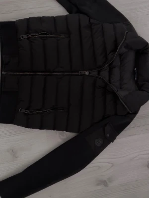 Moncler Cardigan  - Svart pufferjacka med quiltad kropp och släta ärmar. Två sneda dragkedjefickor framtill och ribbade muddar vid midja och ärmslut. Jackan har en modern look och passar perfekt till vintern. Storlek 0 vilket motsvarar XS-S i vanliga storlekar.