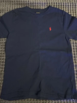 Marinblå t-shirt från Polo Ralph Lauren - Klassisk marinblå t-shirt från Polo Ralph Lauren 10-12 år/ 155-165 cm.med röd broderad logga på bröstet. T-shirten har rund halsringning och korta ärmar. Perfekt basplagg med stilren design och skön passform.