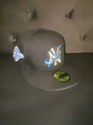 New Era Yankees keps med blå blommor - Snygg svart New Era 59FIFTY keps med New York Yankees-logga framtill dekorerad med broderade blå blommor. Kepsen har platt skärm och patch på sidan. Perfekt för dig som vill sticka ut med en unik och trendig accessoar.