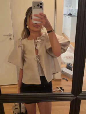 Beige blus med puffärm och knyt - Trendig beige blus med stora puffärmar och två knyt framtill. Blusen har en boxig passform och är tillverkad i ett slätt, lätt glansigt material som ger en lyxig känsla. Perfekt att styla både med kjol och jeans för en cool look.