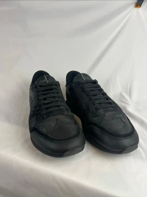 Valentino Rockrunners  - Snygga helsvarta sneakers från Valentino Garavani med diskret logga på plösen. Skorna har en klassisk låg siluett, svarta skosnören och ovandel i mocka och läder. Perfekta för dig som gillar stilrena och exklusiva sneakers. Storlek 44 och i väldigt fint skick.