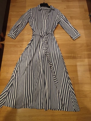 Randig långklänning med bälte - Snygg långklänning med marinblå och vita ränder, knappar framtill och matchande bälte i midjan. Klänningen har krage och långa ärmar med uppvikta muddar. Perfekt för en trendig och avslappnad look.