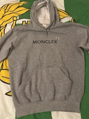 Grå Moncler hoodie  - Snygg grå hoodie från Moncler med svart tryckt logga och klassisk Moncler-patch på bröstet. Hoodien har huva med dragsko, magficka och ribbade muddar. Tillverkad i mjukt bomullsmaterial som är skönt mot huden. Perfekt för en avslappnad och trendig look. 