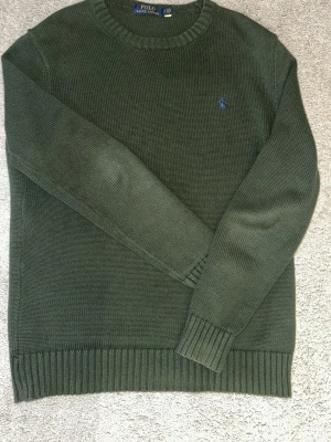 Ralph lauren stickad tröja slimfit  - Slim fit Mörkgrön stickad tröja från Polo Ralph Lauren med rund halsringning och diskret blå logga broderad på bröstet. Tröjan har ribbade muddar vid ärmslut och nederkant. Klassisk och enkel design som passar till mycket.