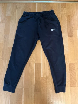 Svarta mjukisbyxor från Nike - Säljer ett par svarta mjukisbyxor från Nike med dragsko i midjan och resår vid bensluten. Byxorna har två sidofickor och en vit Nike-logga på vänster lår. Perfekta för chill eller träning, riktigt sköna och stilrena.