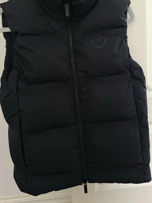 Svart dunväst Knowledge Cotton - Snygg svart dunväst från Knowledge Cotton Apparel med hög krage och dragkedja framtill. Västen har diskret logga på bröstet och är tillverkad i ett mjukt, vadderat material som håller värmen. Perfekt för lager på lager och streetwear-stil.