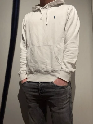 Vit hoodie från Polo Ralph Lauren - Säljer en stilren vit hoodie från Polo Ralph Lauren med klassisk huva och dragsko. Tröjan har en känguruficka framtill och den ikoniska lilla blå loggan broderad på bröstet. Perfekt för en avslappnad och snygg look.