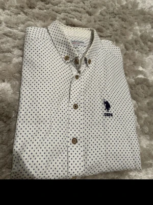 Vit mönstrad skjorta från U.S. Polo Assn. - Snygg vit skjorta från U.S. Polo Assn. med små blå prickar och klassisk krage. Skjortan har knappar framtill och ett broderat blått logomärke på bröstet. Tillverkad i bomull och har lång ärm för en clean och stilren look.