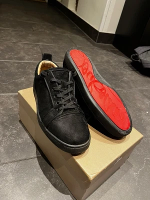 Svarta sneakers från Loouuobs  - Snygga svarta sneakers i mocka från Christian Luuooubs med klassisk röd sula och svarta skosnören. Låg modell med diskret logga på sidan av sulan och bekväm innersula i beige. Perfekta för dig som vill sticka ut med exklusiv stil.