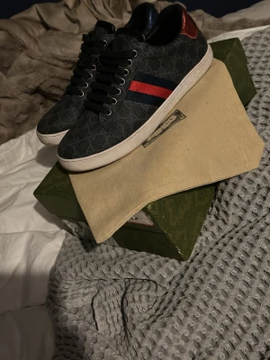 Gucci sneakers med monogram och röd detalj - Snygga sneakers från Gucci i svart med klassiskt GG-monogram över hela skon. Skorna har svarta skosnören, vit sula och de ikoniska röd-blå ränderna på sidan samt en röd metallic-detalj på hälen. Materialet är en mix av canvas och läder.kommer med endast dustbag jag tycker att de är osköna vid tån därför säljer jag dem