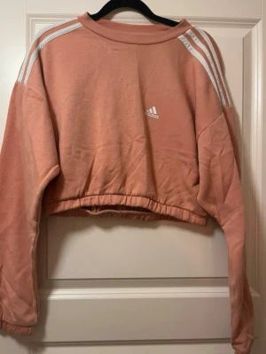 Rosa croppad hoodie från Adidas - Säljer en snygg croppad hoodie från Adidas i en mjuk rosa färg. Hoodien har vita ränder på axlarna och Adidas-logga framtill. Tillverkad i ett skönt bomullsmaterial och har långa ärmar med resår vid midjan för en relaxed vibe.